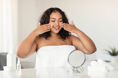 Young black oversize woman covered in towel sitting at table, looking at mirror and applying makeup on her cheeks and smiling, enjoying her brand new cosmetics