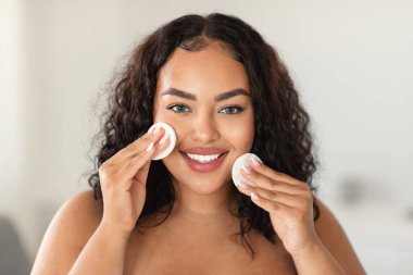 Happy black plus size lady holding cotton pads and smiling at camera, using cleansing lotion or hydrating toner, enjoying face care routine