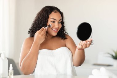 Young black oversize lady making makeup, applying facial powder with cosmetic brush, looking at mirror, copy space. Makeup products and beauty routine concept