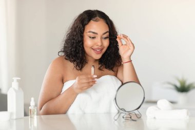 Cosmetics and makeup concept. Happy african american chubby woman looking in mirror and putting mascara on eyelashes in bedroom interior in morning, free space