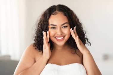 Face massage. African american plus size woman touching her smooth flawless skin on cheeks and smiling at camera, sitting in bedroom interior, wrapped in towel