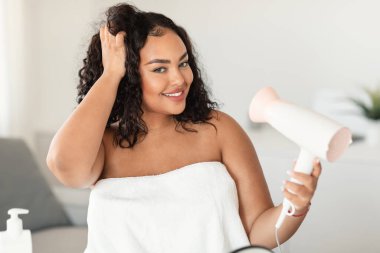 Haircare concept. Happy black plus size lady drying and styling hair with hairdryer, making hairstyle at home. Pretty woman enjoying beauty routine after morning shower
