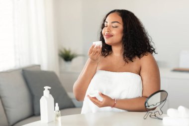 Beauty, cosmetics, skin care concept. Happy black plus size woman holding and smelling cream jar, sitting in bedroom wrapped in towel, free space