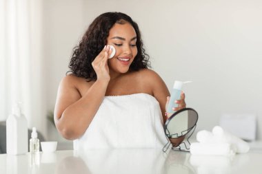 Young black chubby woman using micellar water and cotton pad for skin cleansing at home, sitting in front of mirror, removing makeup from face, copy space