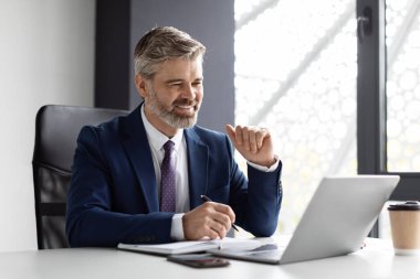 Handsome smiling middle aged businessman using laptop and writing in notepad while sitting at workplace in modern office, successful mature male entrepreneur in suit planning working schedule