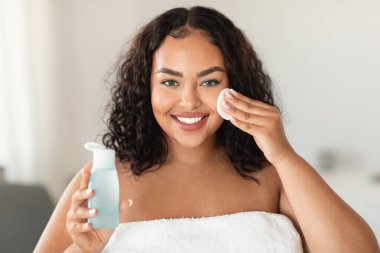 Beautiful black chubby lady cleansing skin with micellar water and cotton pad, making daily beauty routine in bathroom, woman enjoying home skincare treatments