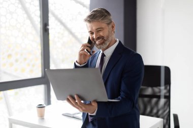 Smiling Mature Male Entrepreneur Talking On Mobile Phone And Using Laptop In Office, Middle Aged Businessman Enjoying Pleasant Phone Conversation With Client And Looking At Computer Screen