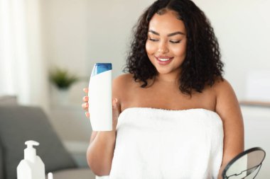 Young black chubby woman advertising shampoo showing bottle and smiling, sitting in bathroom at home, recommending cosmetics. Beauty products concept