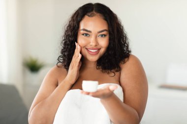 Happy black chubby lady using face cream at home, looking at camera and showing cream jar, applying beauty product on her face, bedroom interior