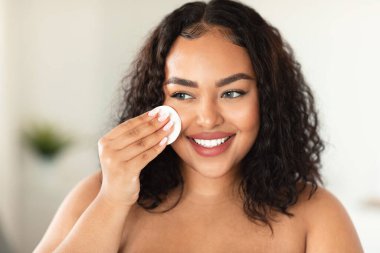 Closeup portrait of black plus size woman using cotton pad, erasing make up from her face, applying facial toner or cleansing milk, bathroom interior, copy space