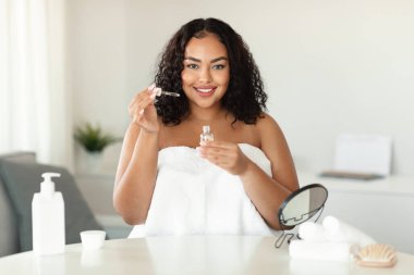 Skincare concept. Plus size black lady applying face serum with dropper while sitting at home, smiling at camera. African american woman moisturizing skin, enjoying self-care routine