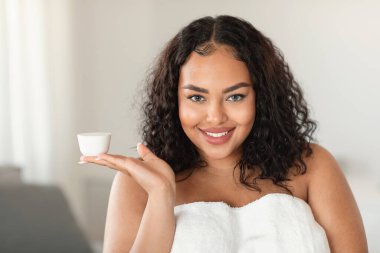 Professional cosmetics for beautiful skin. Beautiful black oversize woman holding jar with moisturising cream on open palm and smiling at camera, standing in bedroom interior