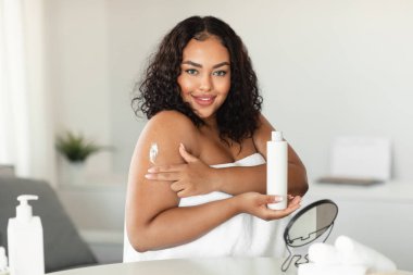 Bodycare and skin nutrition. Happy african american plus size lady using body lotion, holding bottle, doing daily morning routine, pampering herself after bath