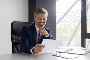 Handsome Mature Male Entrepreneur Reading Financial Papers While Sitting At Desk In Office, Smiling Middle Aged Businessman Checking Annual Reports At Workplace, Enjoying Company Growth, Free Space
