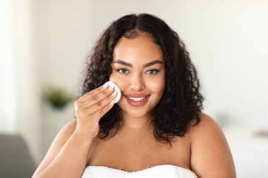 Skincare concept. Happy black overweight lady cleansing face with cotton pad, making self-care beauty routine, standing in light bathroom interior