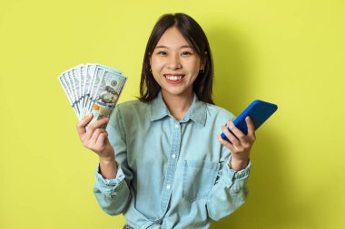 Financial Luck. Cheerful Asian Lady Holding Smartphone And Money Cash Smiling To Camera Standing On Yellow Studio Background. Mobile Banking And Ecommerce Concept