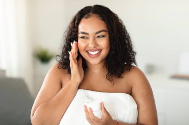 Happy black overweight lady using moisturising face cream, holding jar with beauty product and smiling, enjoying her morning face care routine