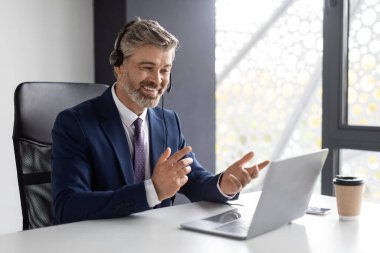 Online Meeting. Smiling mature businessman in headset making video conference via laptop in office, happy middle aged entrepreneur sitting at desk with computer and talking at web camera, copy space