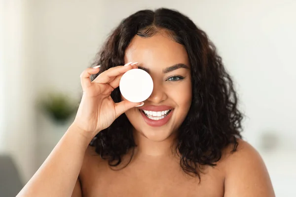 Beauty products. Excited black bodypositive lady holding jar with nourishing cream, covering one eye with new skincare item, standing in bathroom interior