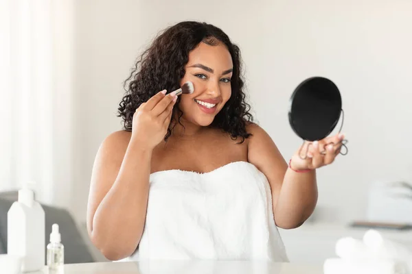 Happy black body positive woman with curly hair in towel looking in mirror and applying powder with brush on face. Basic makeup, routine procedures, cosmetics and beauty care