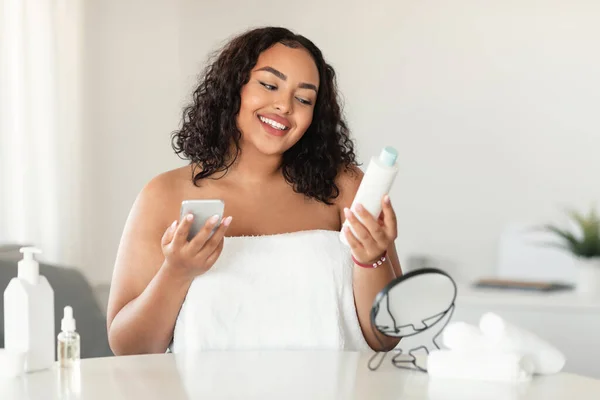 Happy black chubby woman in white towel reading recommendations on lotion bottle and using smartphone, sitting in bedroom interior and smiling