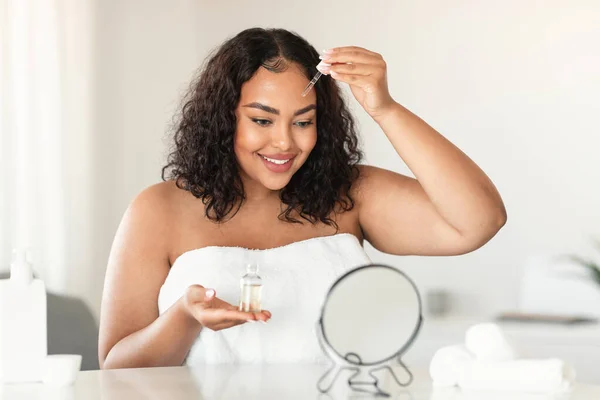 Skincare concept. Pretty black chubby woman applying face serum with dropper, sitting in front of mirror and smiling. African american lady moisturizing skin at home