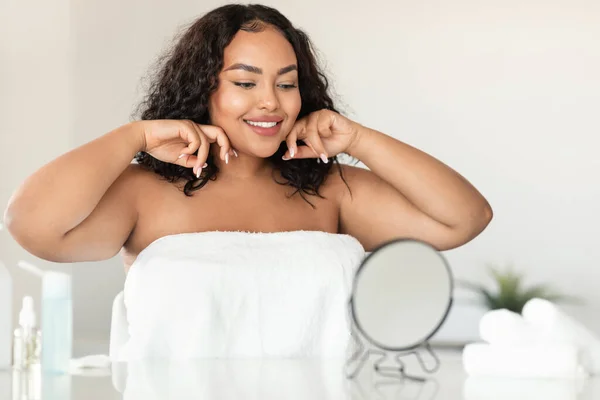 Beautiful black chubby lady making lymphatic drainage self massage, sculpting jaw line in front of mirror, free space. Young woman doing domestic skin care procedure at home
