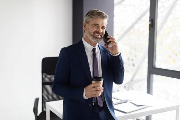 Handsome Middle Aged Man In Suit Talking On Cellphone And Drinking Coffee In Office, Smiling Mature Male Entrepreneur Holding Cup With Takeaway Drink And Enjoying Pleasant Mobile Conversation