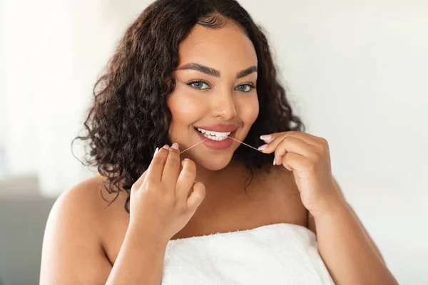 Young black chubby woman cleaning teeth with dental floss at home and looking at camera. Happy plus size lady performing everyday dental care. Oral hygiene, healthy lifestyle concept