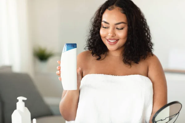 Young black chubby woman advertising shampoo showing bottle and smiling, sitting in bathroom at home, recommending cosmetics. Beauty products concept