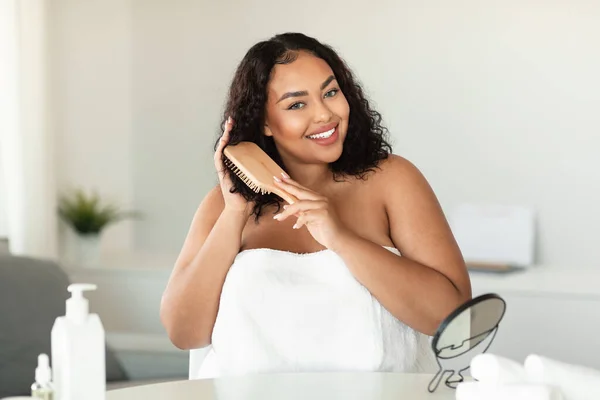 Happy black chubby woman brushing her curly hair with comb and smiling at camera, african american lady wrapped in towel making haircare routine at home