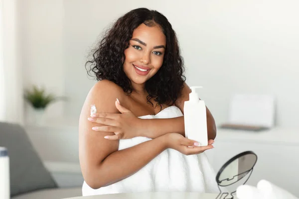 Young black chubby lady applying moisturizing body lotion on shoulder, holding bottle and smiling at camera, woman using nourishing cream for skin, making beauty treatments at home
