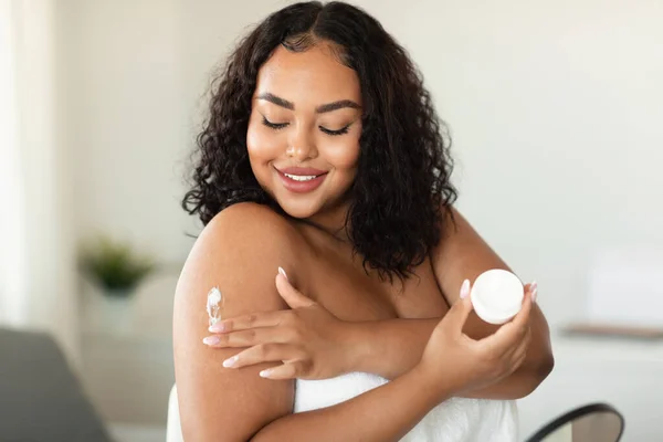 Young african american chubby woman applying skin butter or mosturizer on her shoulder after morning shower and smiling, holding jar with cream