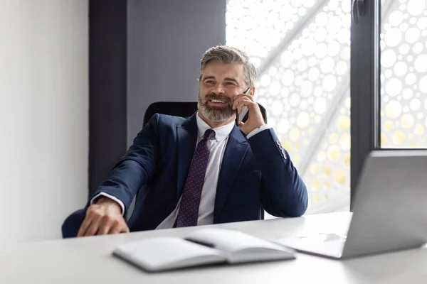 Phone Call. Smiling Mature Businessman In Suit Talking On Cellphone At Workplace, Handsome Relaxed Male Entrepreneur Sitting At Desk In Office, Enjoying Pleasant Mobile Conversation, Free Space