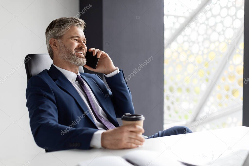 Happy Mature Businessman Having Work Break, Talking On Cellphone And Drinking Takeaway Coffee While Sitting At Desk In Office, Middle Aged Male Entrepreneur Relaxing At Workplace, Copy Space