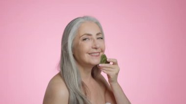 Beauty care procedures. Happy beautiful grey haired senior lady doing skin lifting massage with gouache stone, smiling to camera, pink studio background, slow motion