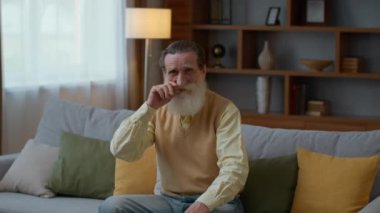 Retirement concept. Handsome senior bearded gentleman resting on sofa at home, smiling to camera and fixing his moustache, slow motion, free space