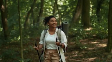 Positive cardio and tourism. Young happy african american woman tourist with backpack walking in forest with sticks, enjoying nature, slow motion, tracking shot
