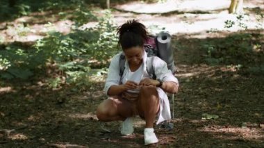 First medical aid in forest. Young positive african american woman hiker putting adhesive band on knee after getting injured, enjoying backpacking trip in forest, slow motion