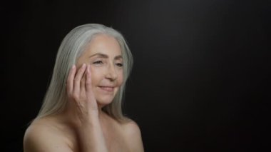 Aging and special cosmetology. Studio portrait of beautiful senior grey haired lady rubbing skin lifting cream on her face, posing with bare shoulders over black background, slow motion, empty space