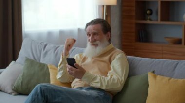 Online win for everyone. Excited bearded senior grandfather web surfing on smartphone, reading message and laughing, enjoying success, sitting at home, tracking shot, slow motion, free space