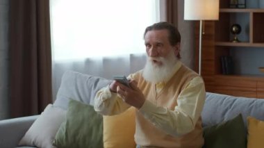 Great retirement savings. Excited senior bearded man throwing dollar banknotes in air, sitting on sofa at home, enjoying win or long awaited payment, slow motion