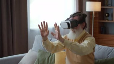 Modern entertainment devices. Happy senior bearded man using VR headset at home, enjoying futuristic game, pushing invisible buttons, tracking shot, slow motion