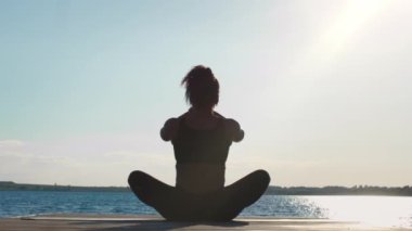 Unity with nature. Back view shot of peaceful black woman meditating outdoors, sitting on wooden pier near big water in morning, slow motion, free space