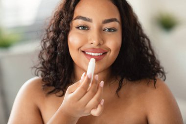 Portrait of black body positive lady applying lip balm moisturizing skin, looking at camera. Lips skincare and makeup, self-care cosmetics advertisement concept