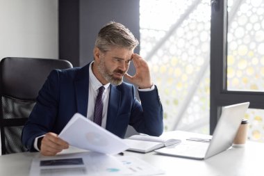 Focused Middle Aged Businessman Working With Documents And Laptop In Office, Stressed Mature Male Entrepreneur Using Computer At Workplace And Checking Company Financial Papers, Copy Space