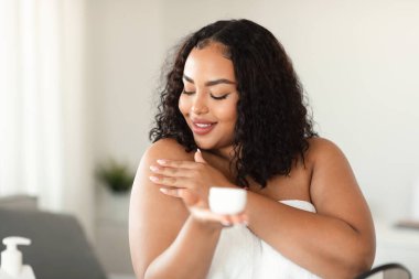 Black plus size lady applying body cream on shoulder after shower in morning at home. Happy african american woman making beauty procedure, pampering herself indoors