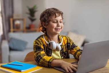 Modern education for youngest generation. Positive little schoolboy typing on laptop, doing home task or watching online lesson, sitting at desk at home, free space