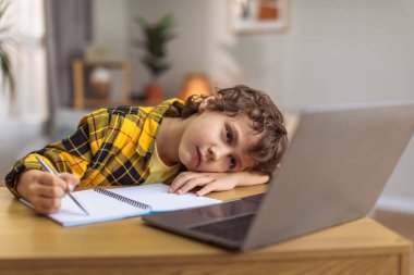 Boring upset little boy lying on hand and writing in copybook, watching lesson online on laptop at home, empty space