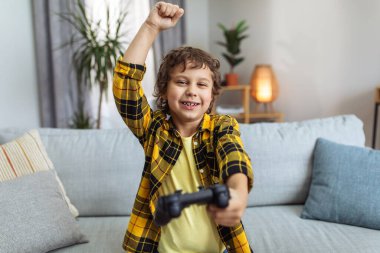 Online win. Happy little boy gamer with joystick celebrating victory in video game, laughing and gesturing to camera, sitting at home, close up portrait